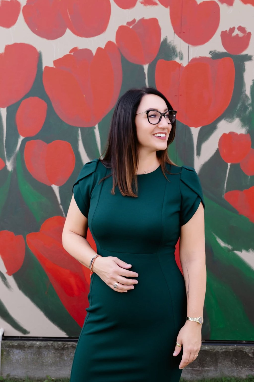 A woman with long dark hair and glasses in a green dress stands in front of a wall adorned with red tulips.
